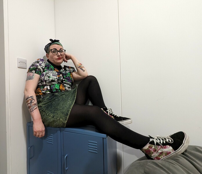 woman with green hair sitting on a metal blue cabinet, wearing a rock shirt, green dyed denim skirt, black tights and black and flower vans trainers