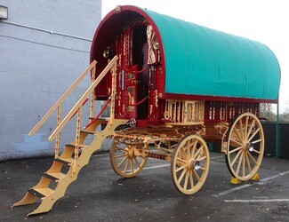 A red and yellow traditional Gypsy caravan with a green bow - top roof. Its steps are down and it is parked in the carpark outside a community centre and school.