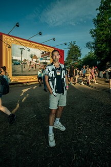 young Roman male wearing beige shorts, black top and grey patterend shirt. Standing with his hands in his pocket in front of billboard design among young crowd