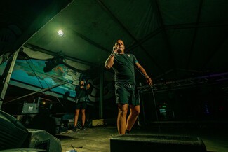 Tall white male wearing a black t-shirt and black shorts standing on a stage with a mic in his hand.