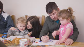 Two Adults sat at table with children on their laps writing 