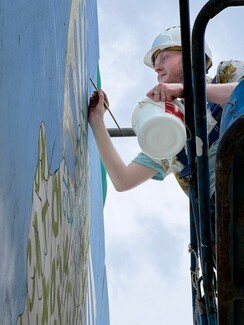 white male painting on platform holding paint bucket and brush