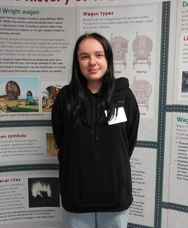 A white young woman stands in front of a display about traditional Gypsy caravans. She has long, straight black hair and dark eyes. She is wearing a black hoodie, light blue jeans and a white name tag. She is smiling.