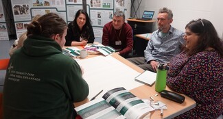 Six people are sat around a table talking. They are: a white man with long brown hair in a bun, a short beard and a green hoodie, a white woman with long brown hair, a floral dress and a pair of glasses perched on her head, a white man with grey hair and short beard and a blue floral shirt, a white man with grey hair, a brown jumper and a colourful lanyard, a white woman with long black hair, a black cardigan and a white name tag and a white woman with long blonde hair and a brown cardigan.