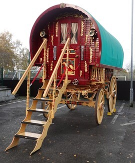 A red and yellow traditional Gypsy caravan with a green bow - top roof. Its steps are down and it is parked in the carpark outside a community centre and school.