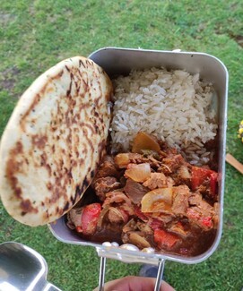 Against a background of grass, a hand holds a camping pan with game curry, rice and a naan bread in it.