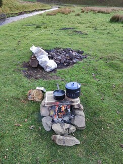 In a grass field, with a stream in the background, there is an open fire surrounded by rocks. On it sits a metal grate with a black kettle and pa n on top of it. A dead rabbit sits next to the fire.