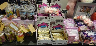 In front of a black background, a table h olds various boxes filled with packaged vegan marshmallows . There are also several miniature clear sweet jars , filled with brightly coloured vegan sweets