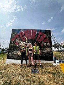 Three workers posing infront of billboard. The billboard has two people riding horses and in bid red font days, "Atchin Tan."