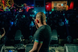 A white male, Sugar Shane, is on a stage facing his audience. The dark crowd is lit by LED lights, blue, yellow, and red