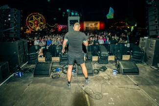 A white male, Sugar Shane, stands on the edge of a stage performing to an unseen crowd. A red and yellow lit ferris wheel is in the background.