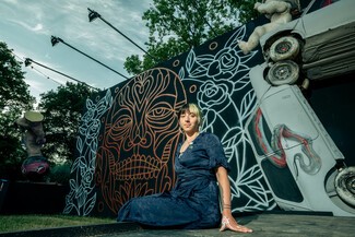young women wearing a navy velvet patterned dress sitting on a stage with neon spray paint artwork and metal car sculpture behind her.
