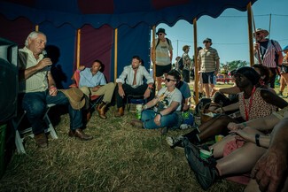 Medium size crowd gathers in blue and red circus tent looking at speaker on the left. He is speaking into the mic and sat next to two male speakers.